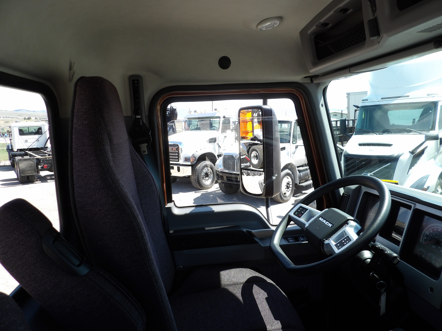 Interior cockpit of a 2026 Mack MD7 truck showing steering wheel and driver seat with dashboard and side window. visible outside are other trucks.