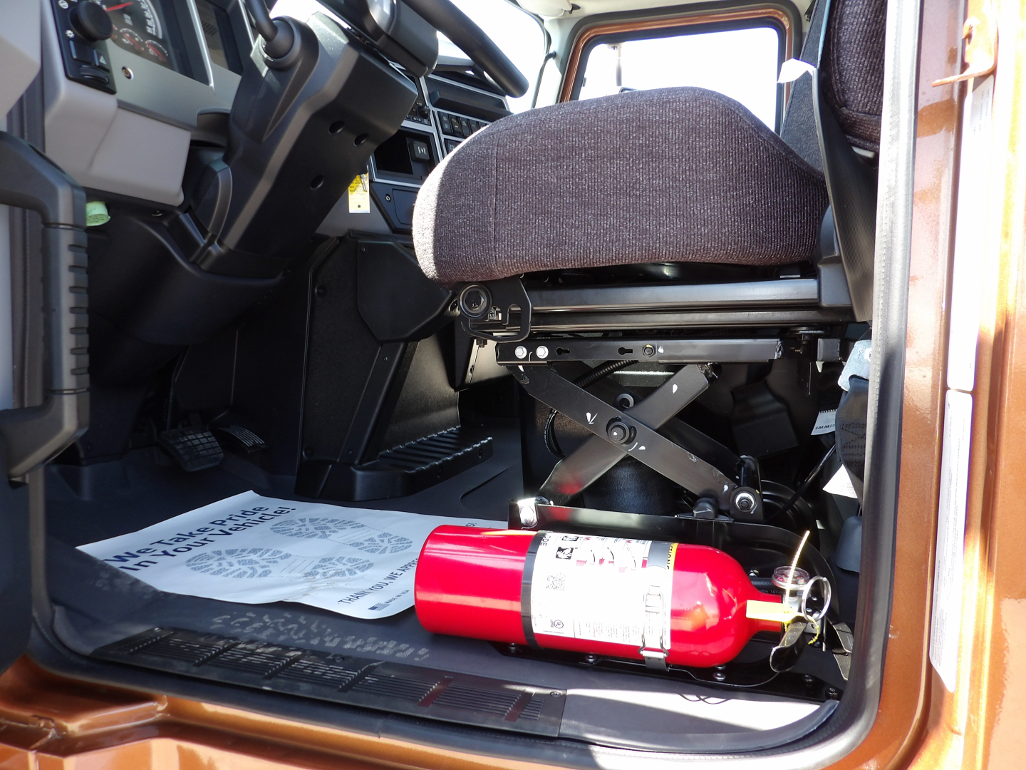 Truck cockpit interior with driver seat visible, control pedals, and a red fire extinguisher on the floor near the door sill.
