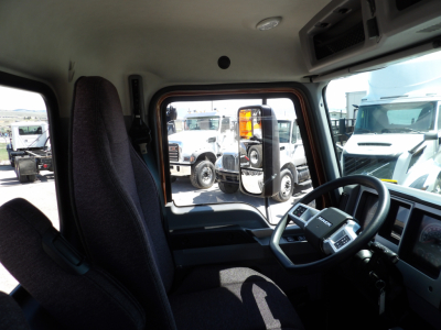 Interior cockpit of a 2026 Mack MD7 truck showing steering wheel and driver seat with dashboard and side window. visible outside are other trucks.