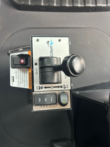 Mid-connector joystick control panel labeled APPSCQ? with safety warnings, buttons labeled COG, HORN, and a small red emergency switch; a gray joystick with black knob; warning label and switch housing visible.