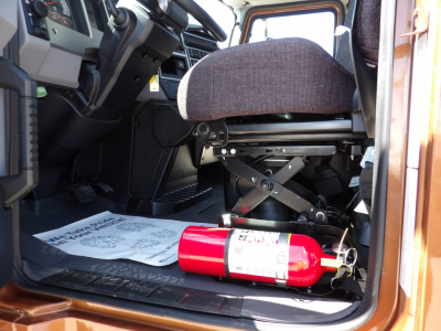 Truck cockpit interior with driver seat visible, control pedals, and a red fire extinguisher on the floor near the door sill.
