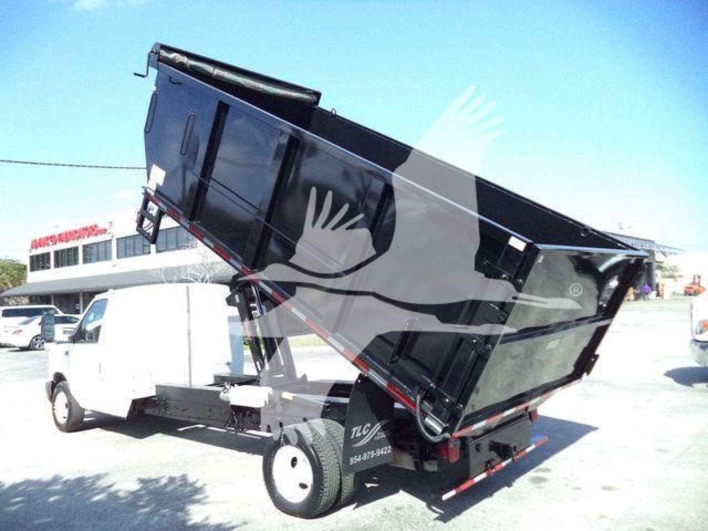 Truck dump bed raised on a Ford E-450 with dump body in operation, white cab, black steel dump box, rear safety lights visible.