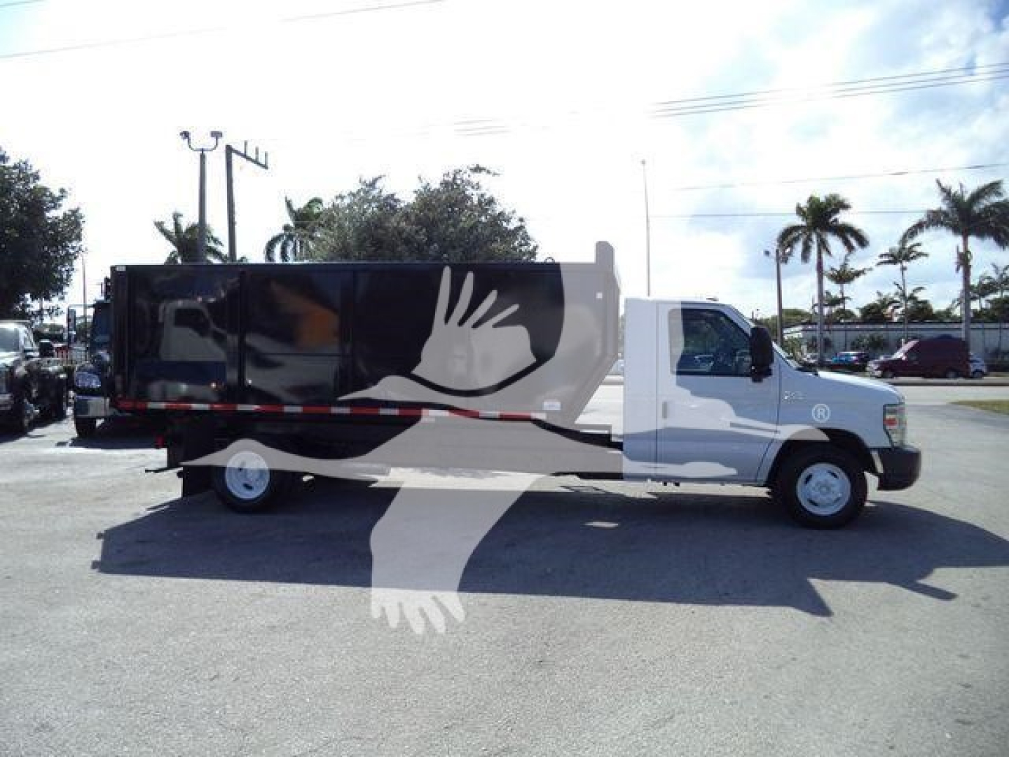 Ford E450 dump truck side view with black and white graphic wrap featuring large hands; side mirror and cab visible; palm tree scene in background.