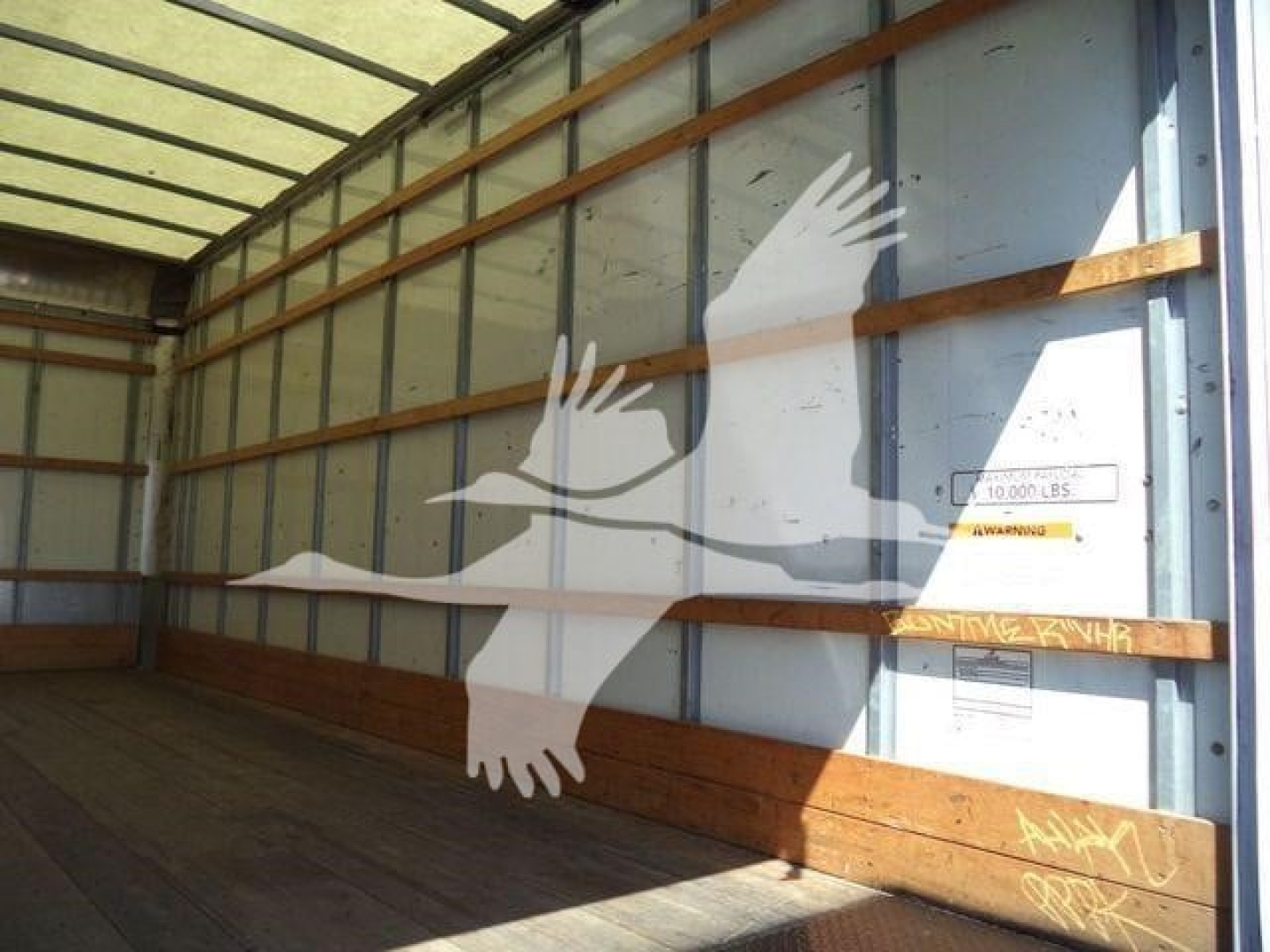 Truck cargo area interior with side wall and wooden rails; faint labels on the wall include a small yellow warning sticker and a silver plate; large white bird logo silhouette overlays the image.