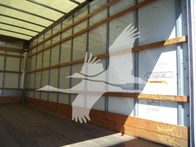 Truck cargo area interior with side wall and wooden rails; faint labels on the wall include a small yellow warning sticker and a silver plate; large white bird logo silhouette overlays the image.