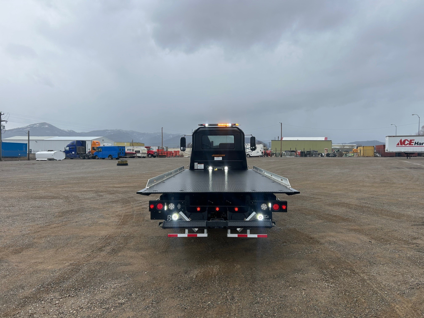 Rear view of a new Mack MD6 rollback tow truck in a lot, with flatbed, warning lights on top, and license-plate area. Visible text: none clearly legible.