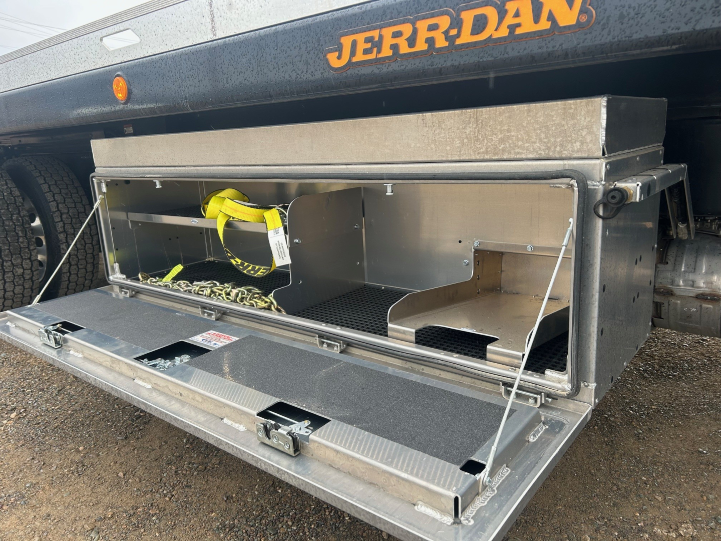 Truck bed tool/storage box mounted under rear of a Mack MD6 rollback truck, open with metal storage compartment and yellow strap inside.