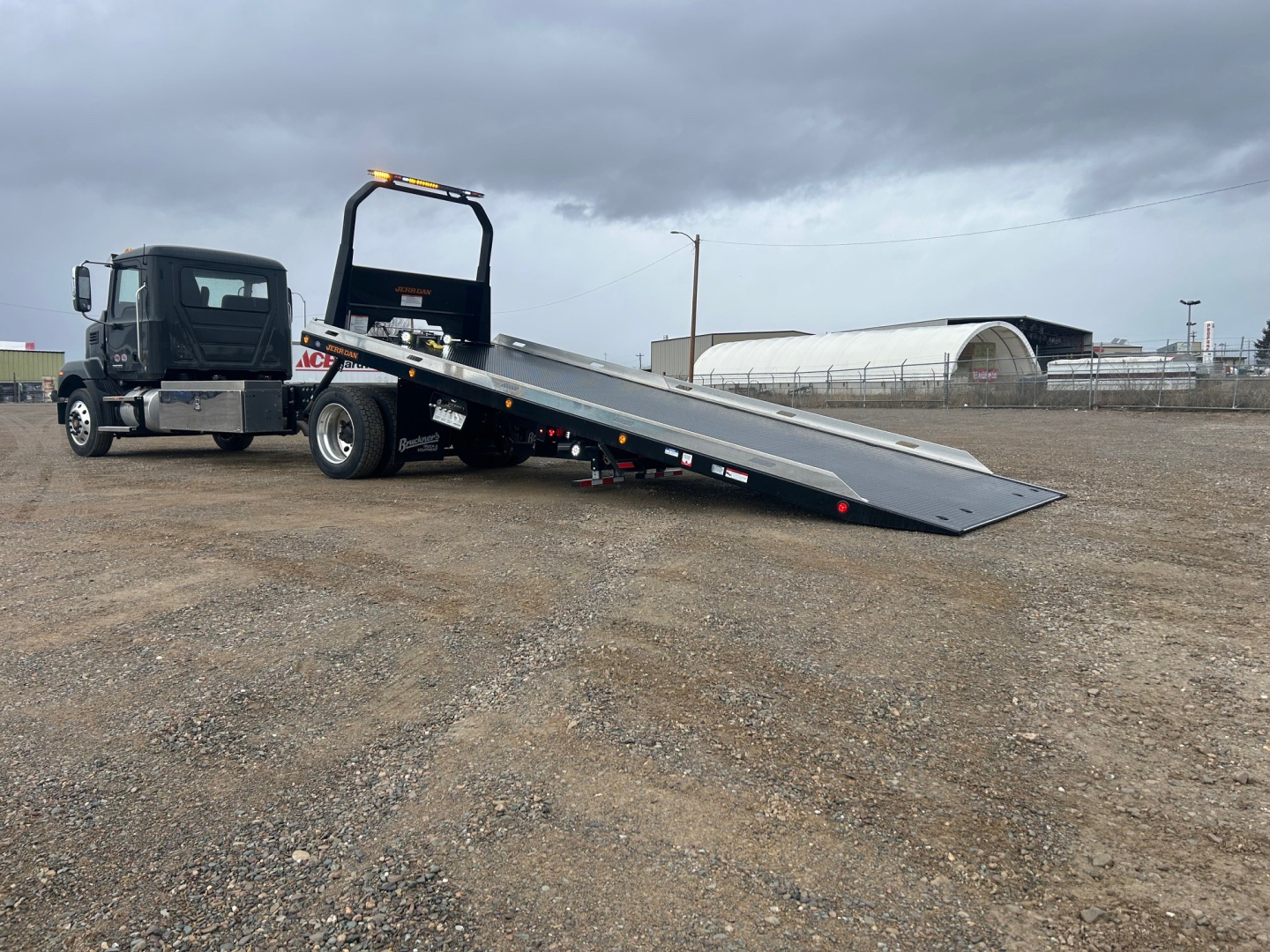 Tow truck with hydraulic rollback tilt bed, black cab, low-angle view, in an open lot.