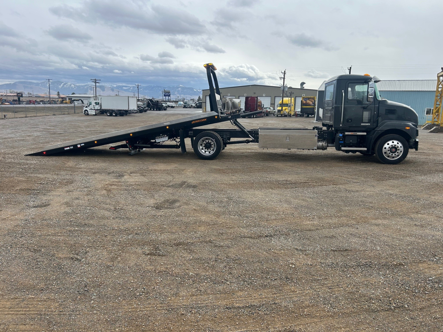 Truck cab with a long wheelbase rollback flatbed, side view showing hydraulic tilt bed and chain/tower equipment in an industrial yard.