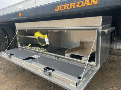 Truck bed tool/storage box mounted under rear of a Mack MD6 rollback truck, open with metal storage compartment and yellow strap inside.