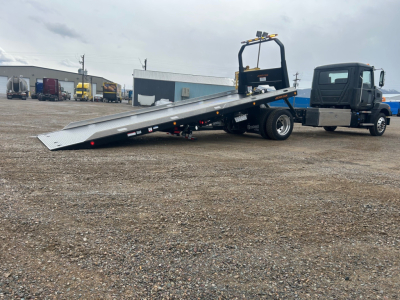 Tractor unit with a lowered aluminum tilt-bed rollback tow truck, cab on right, metal ramp extended to the ground.