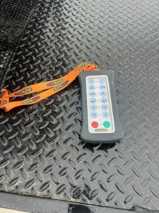 Control remote with blue and gray keypad on a black diamond plate surface. Orange lanyard attached. Buttons labeled with numeric or function icons.