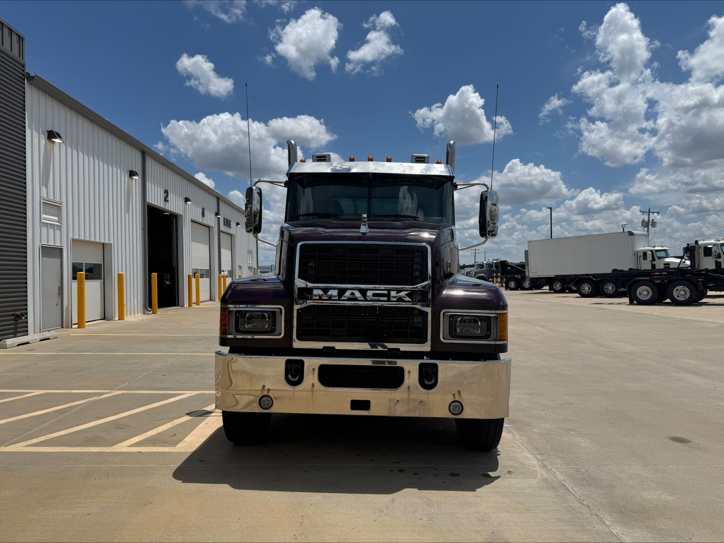 Mack PI64T conventional daycab front view with black grille and chrome bumper, large Mack badge, clear windshield, parking lot with other trucks in background.