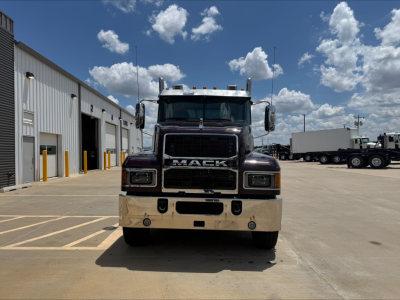 Mack PI64T conventional daycab front view with black grille and chrome bumper, large Mack badge, clear windshield, parking lot with other trucks in background.