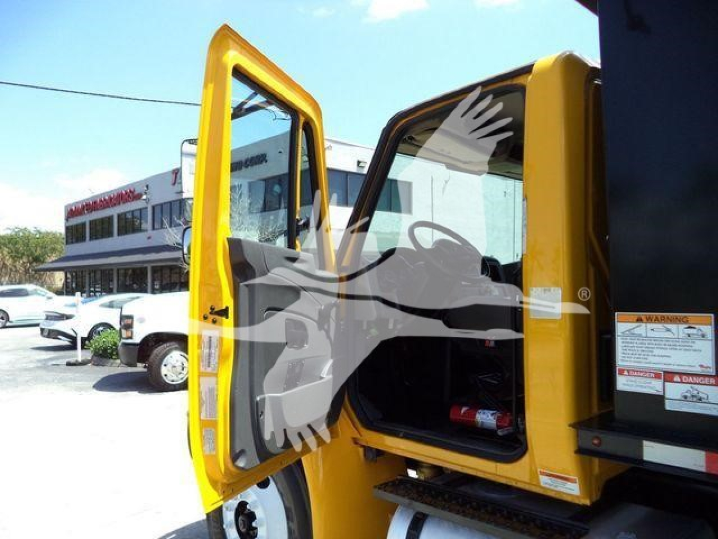 2019 International 4300 truck cab with open yellow door, showing interior and warning labels on the door frame. In background a dealership lot with another truck.