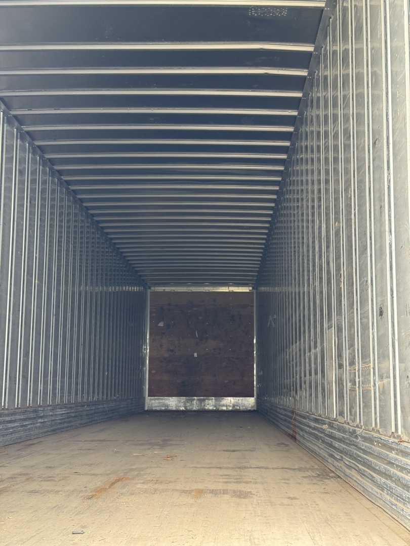 Trailer trailer interior view with metal sidewalls and wooden back wall, empty cargo space, corrugated ceiling, lighting from front opening.