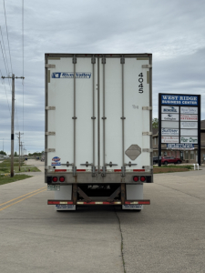 Rear view of a white utility trailer with doors closed, number 4045 on right door, Rivers Valley logo stickers, and bumper reflectors. Street and signboards in background.