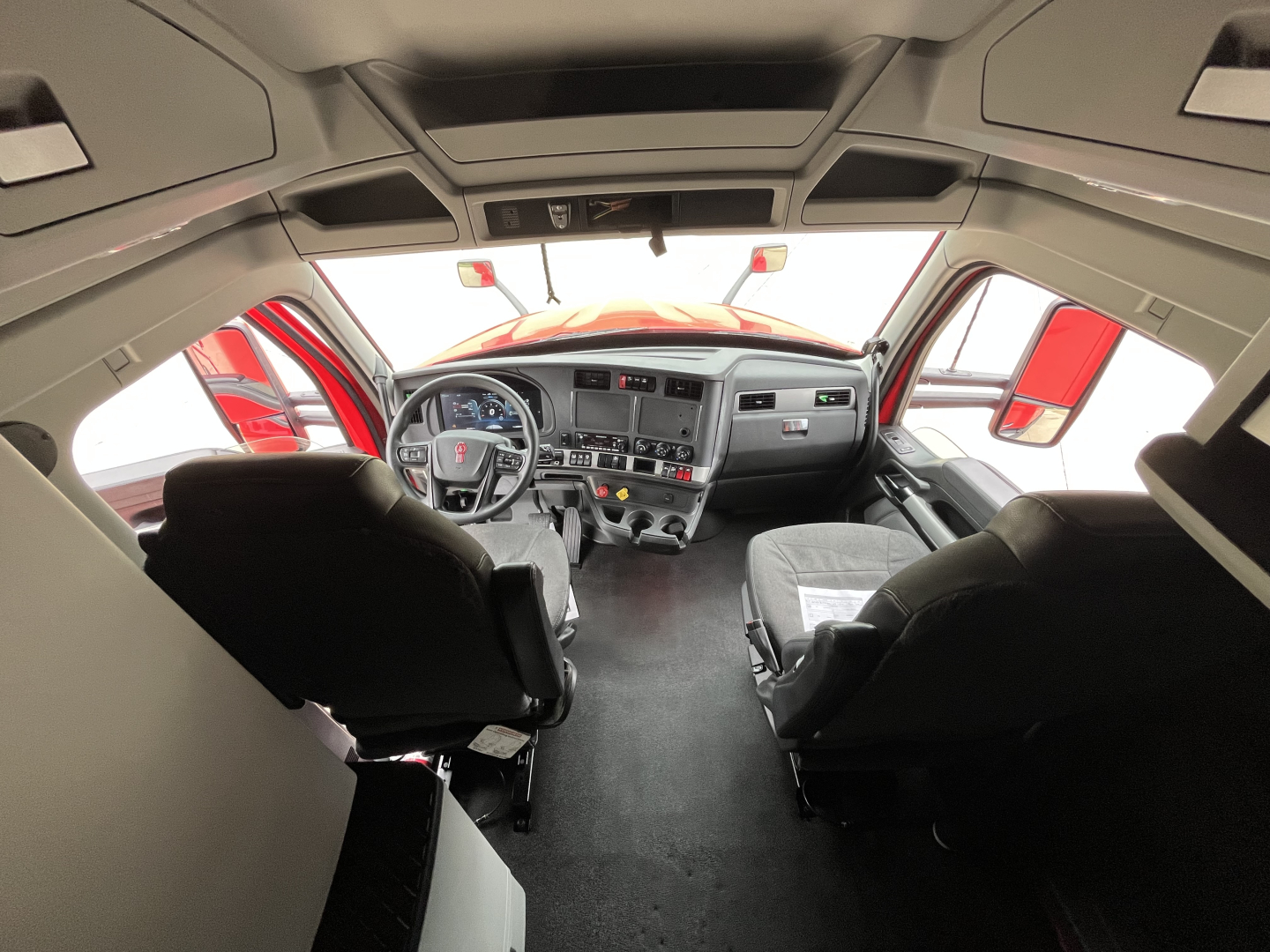 Kenworth T680 cab interior view from ceiling aft shows dashboard, steering wheel, driver and passenger seats, center console, and large windshield with red exterior mirror accents.