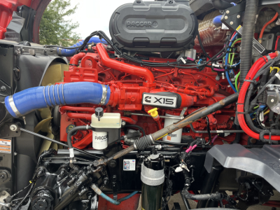Kenworth T680 engine bay with bright red engine and blue intercooler hose, showing X15 badge on valve cover and white reservoir, wires and components visible.