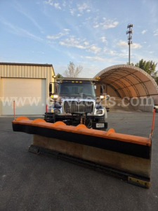 Truck with a large orange snow plow blade front and center, International WorkStar 7600, showroom lot background, stock number visible, VIN plate may be on dashboard.