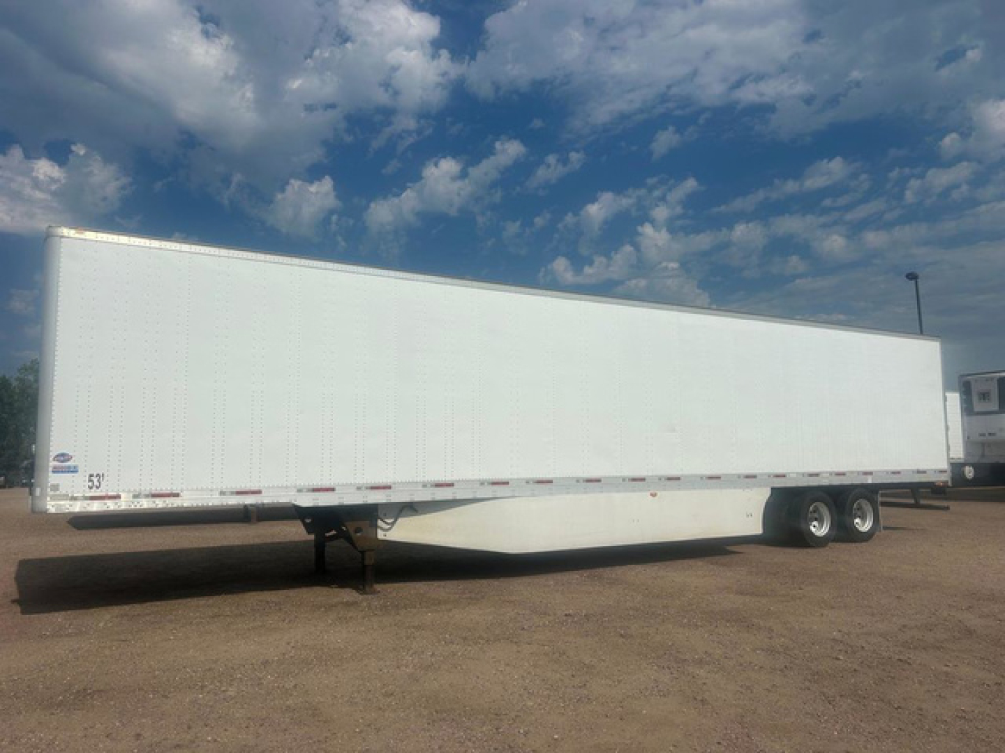 Box trailer extending on a gravel lot under a blue sky showing white cargo trailer and tandem axle wheels.