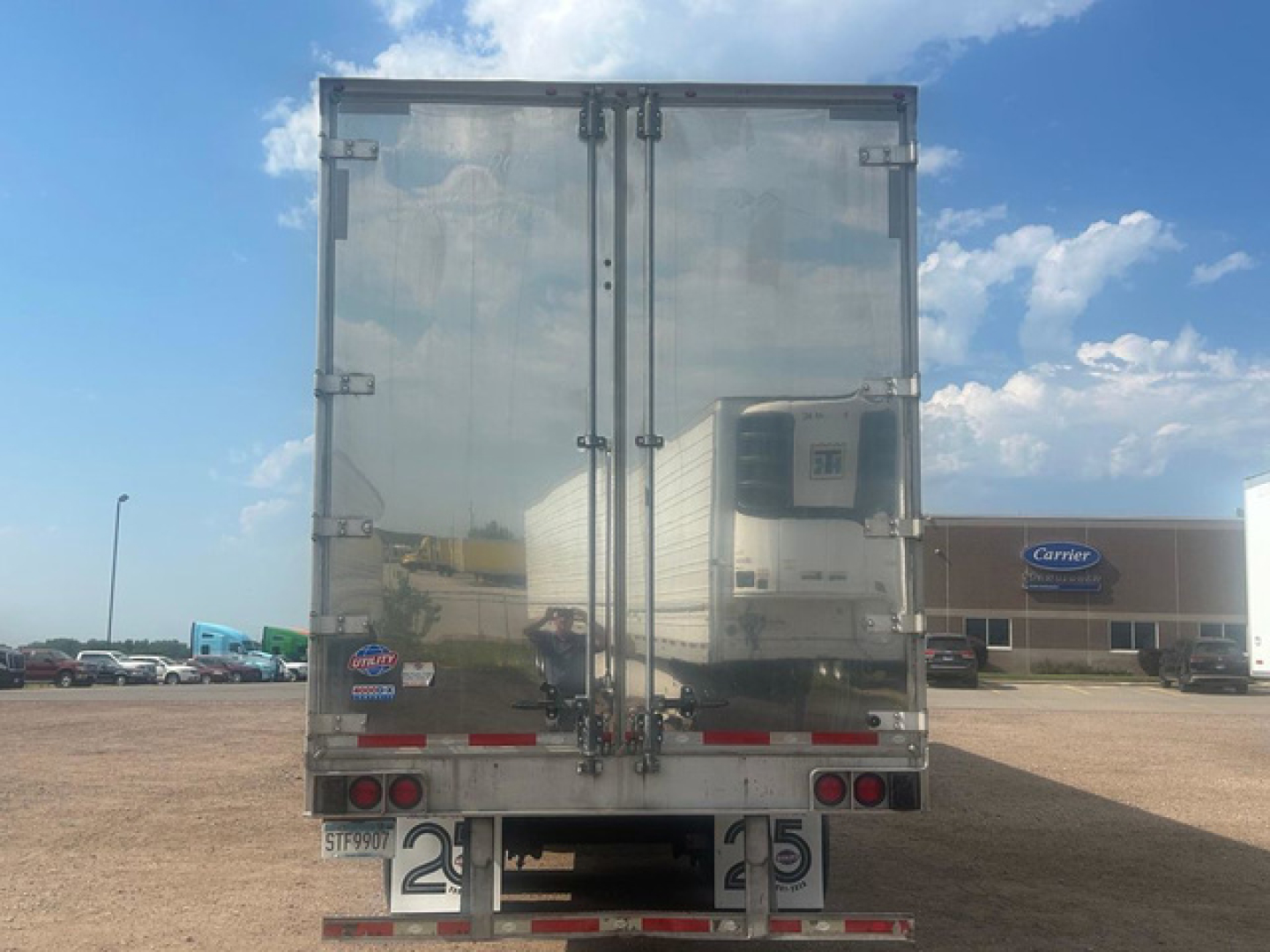 Rear view of a reflective utility van trailer with double doors, taillights, bumper, and Carrier building in the background.