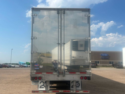 Rear view of a reflective utility van trailer with double doors, taillights, bumper, and Carrier building in the background.