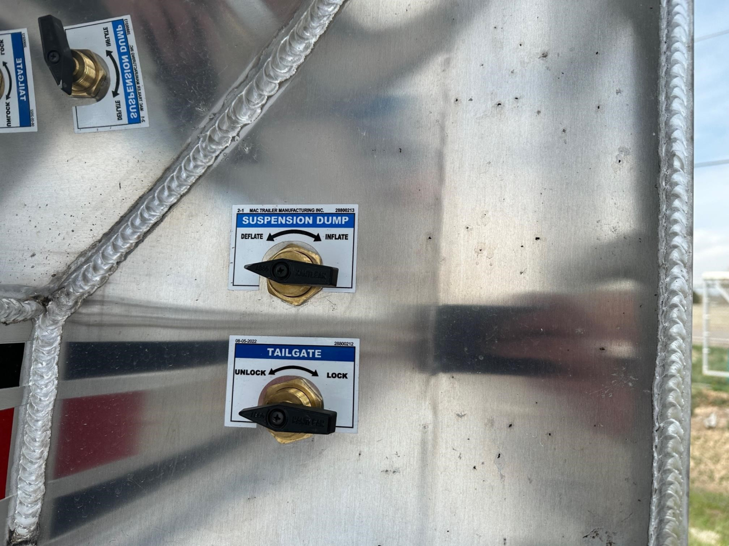 Utility panel on a dump trailer showing labeled control switches: Suspension Dump with INFLATE/DEFLATE levers and Tailgate with UNLOCK/LOCK levers; aluminum body with braided hoses.