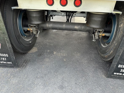 Trailer rear undercarriage showing tandem axles and suspension; round end dump trailer with black steel framework and blue wheels; hydraulic lines and leaf springs visible.