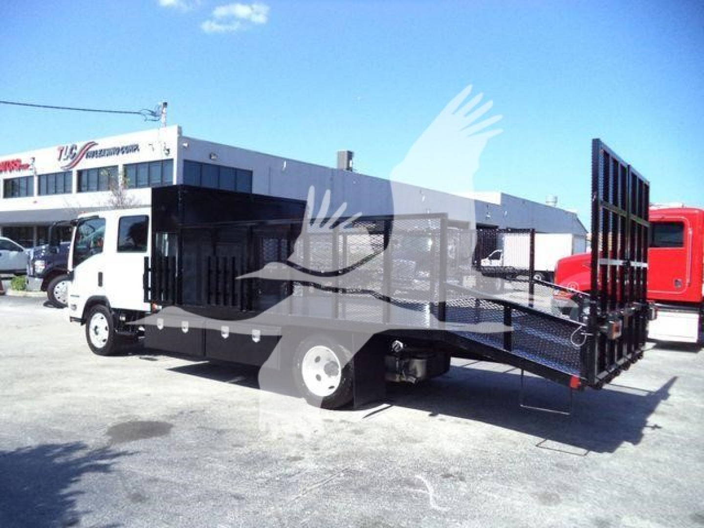 Isuzu NPR-HD landscape truck with flatbed stake bed and hydraulic ramp, in a dealership lot.