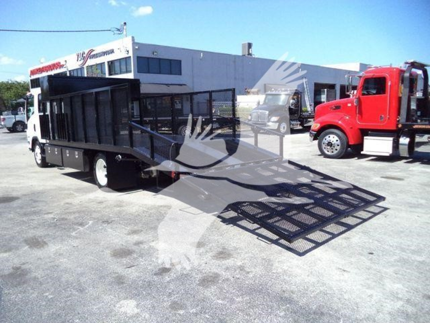 Isuzu NPR HD landscape truck with a ramp and flatbed, parked in a lot next to a red truck; operator cab and black metal cargo area visible.