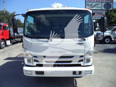 Isuzu NPR-HD truck front view with white cab, large windshield, and grille; visible badges show ISUZU, headlights, and bumper. Background includes other trucks and lot signage.