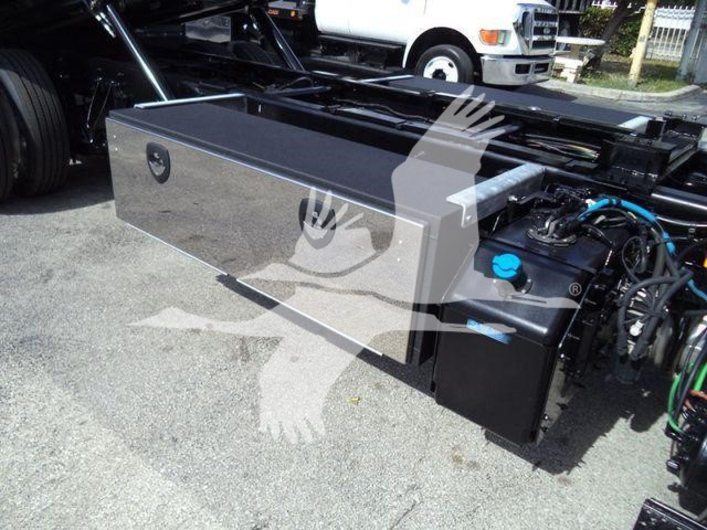 Truck equipment bay showing large rectangular aluminum toolbox with lid and hydraulic hose connections on the right side; blue fluid cap visible, black engine area adjoining.