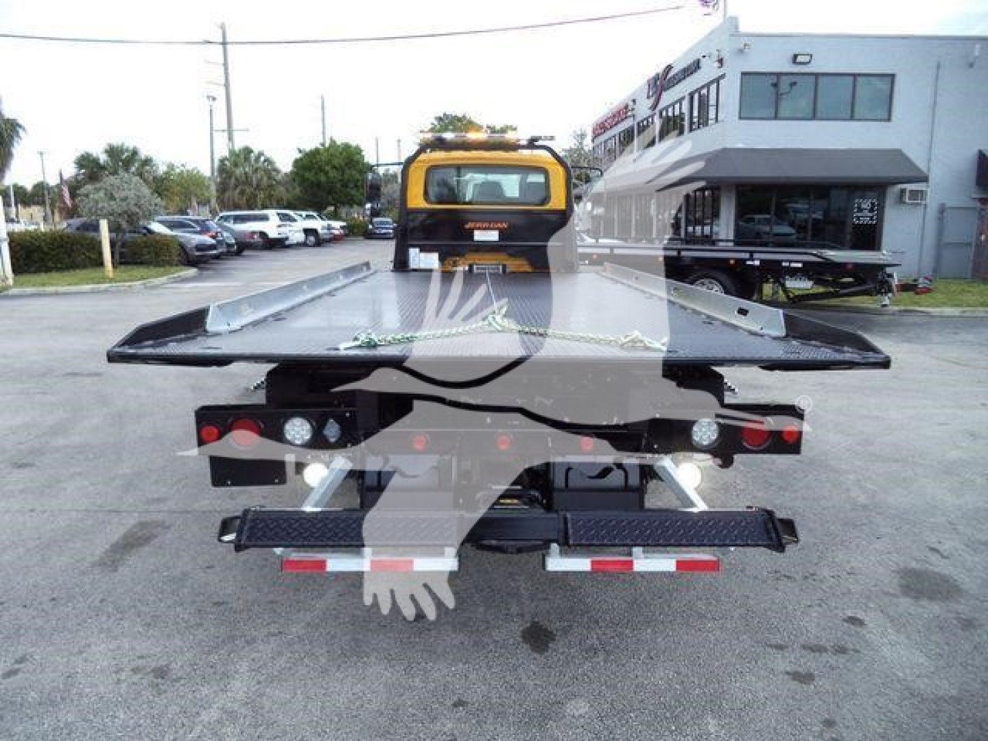 Flatbed tow truck rear with hydraulic winch and wheel chock, yellow cab, dealership building in background.