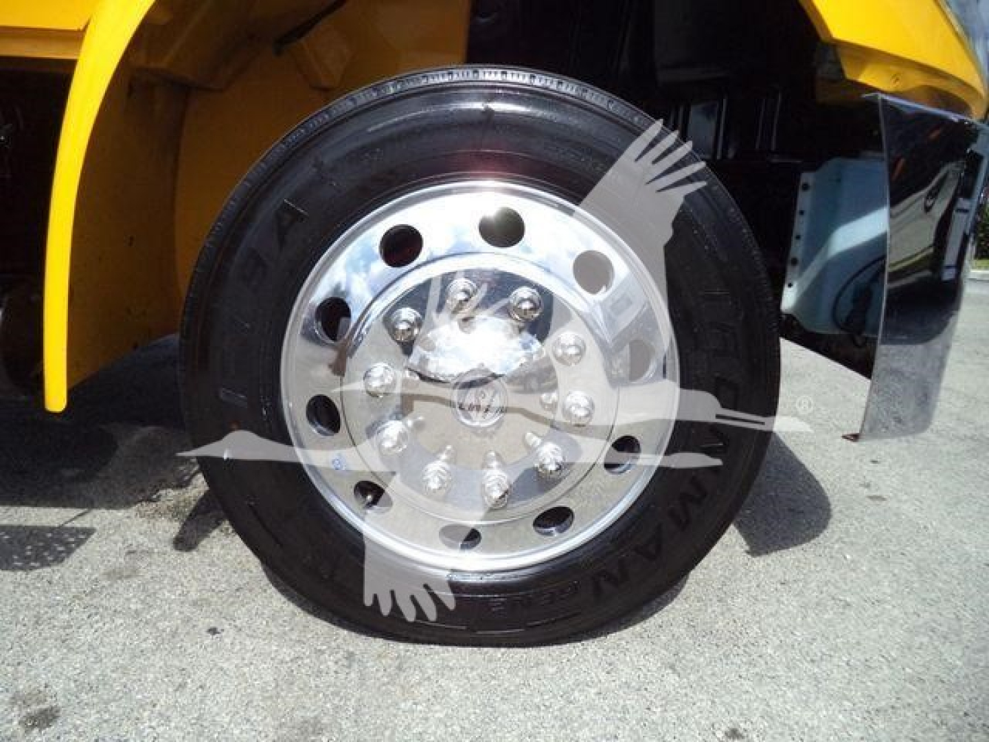 Wheel and tire of a yellow truck with polished steel wheel showing lug nuts; partial fender and mudguard visible.
