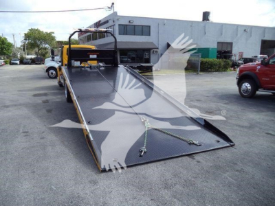 Heavy-duty tow truck with a lowered flatbed in a parking lot; visible are a black diamond-plate steel ramp bed with safety chain and hook, and a small yellow cab in distance.