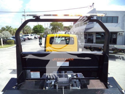 Rear view of a 2019 International 4300 rollback truck bed with its black steel frame and ROPS, hydraulic tow hitch, and platform; visible yellow cab in background and dealership lot.