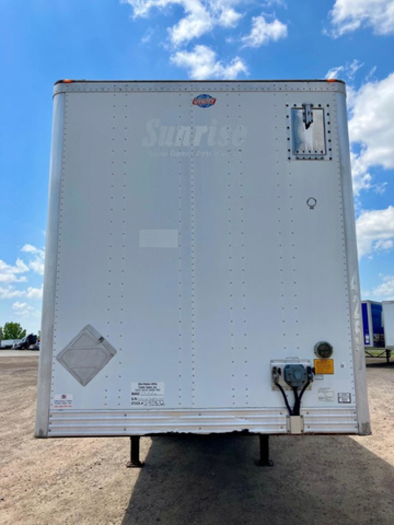 Rear view of a white utility trailer with vertical riveted panels, brand logo and back door latch; small labels and warning decals near bottom left; hitch platform and stand visible.