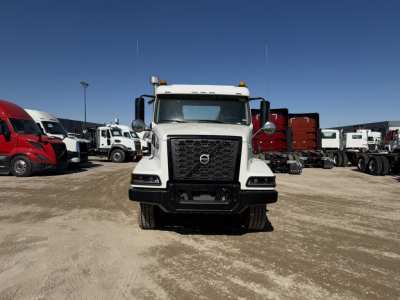 Front view of a white Volvo VHD86B300 cab and chassis truck with black grille, bumper, and large side mirrors. Other trucks parked in the background.