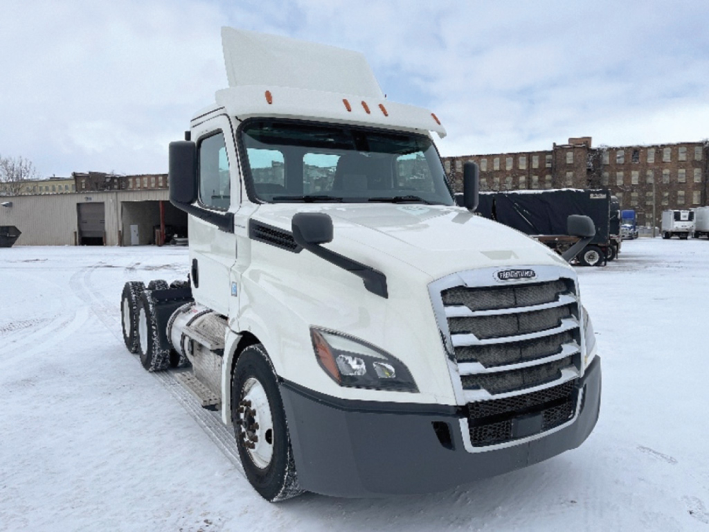 Freightliner Cascadia126 tractor in snowy lot, front-left view showing chrome grille, headlights, large bumper, white cab with raised sleeper, dual rear axles, snow on ground.