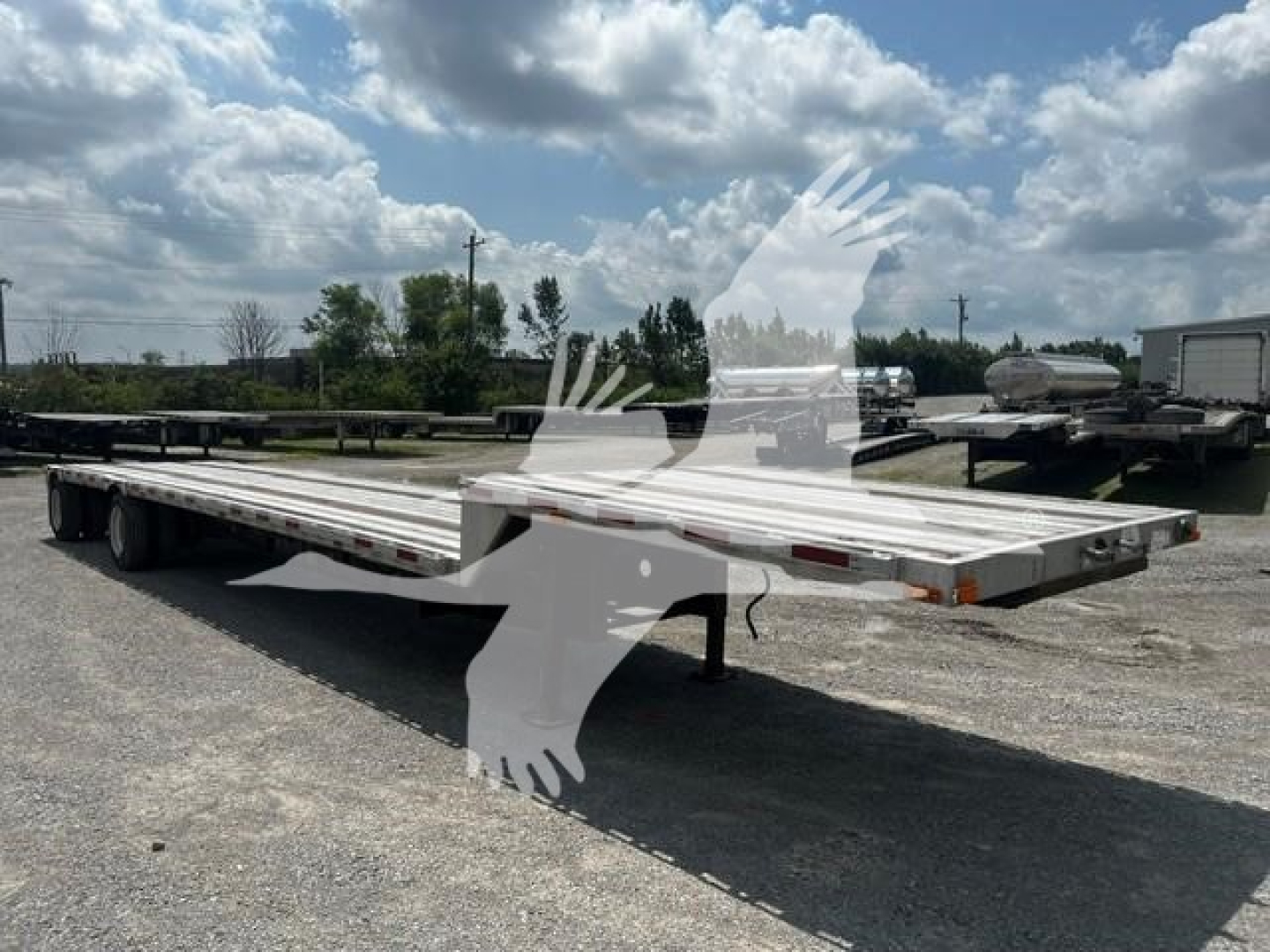 Flatbed trailer with long deck and dual rear axles, empty, in an open lot under blue sky with clouds.