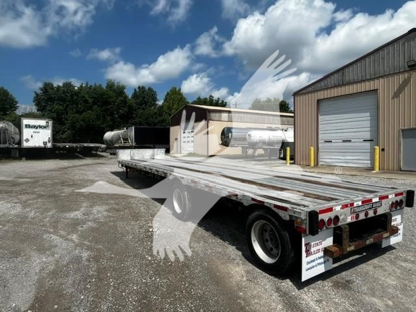 Transcraft DTl-2000 drop deck trailer showing flatbed with rear lights, registration decals, and wheel area; industrial yard with buildings in background.