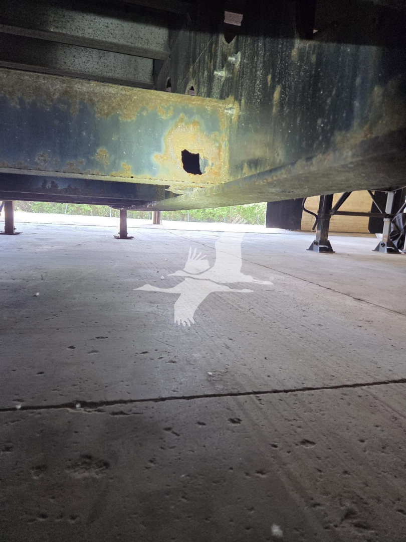 Underneath a Transcraft trailer frame showing a rusted, damaged crossmember and underside structure on a concrete lot.