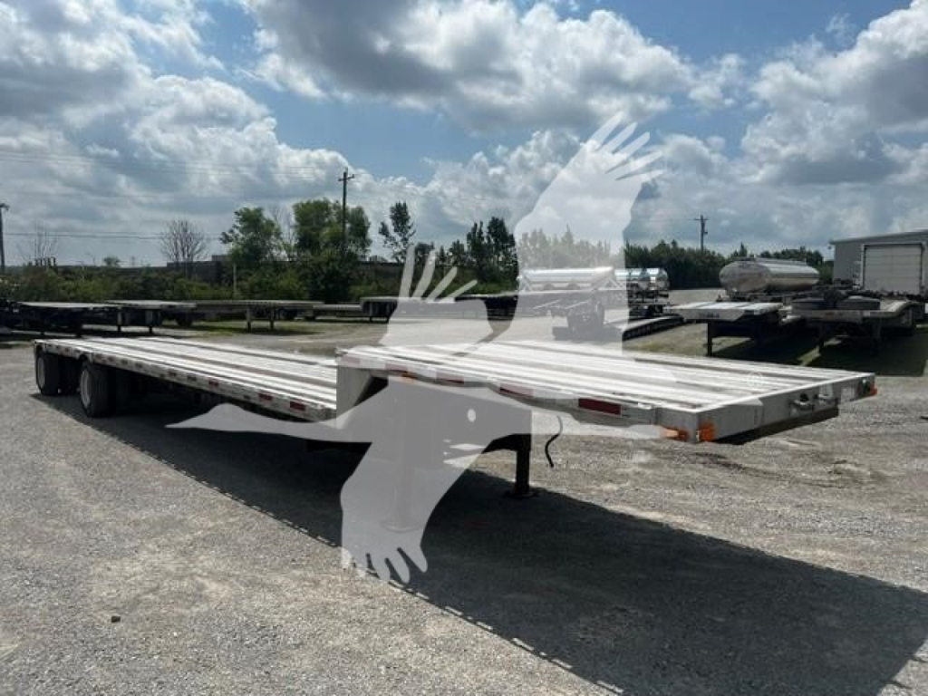 Flatbed trailer with long deck and dual rear axles, empty, in an open lot under blue sky with clouds.