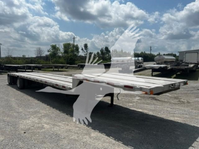Flatbed trailer with long deck and dual rear axles, empty, in an open lot under blue sky with clouds.