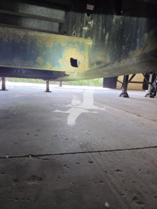 Underneath a Transcraft trailer frame showing a rusted, damaged crossmember and underside structure on a concrete lot.