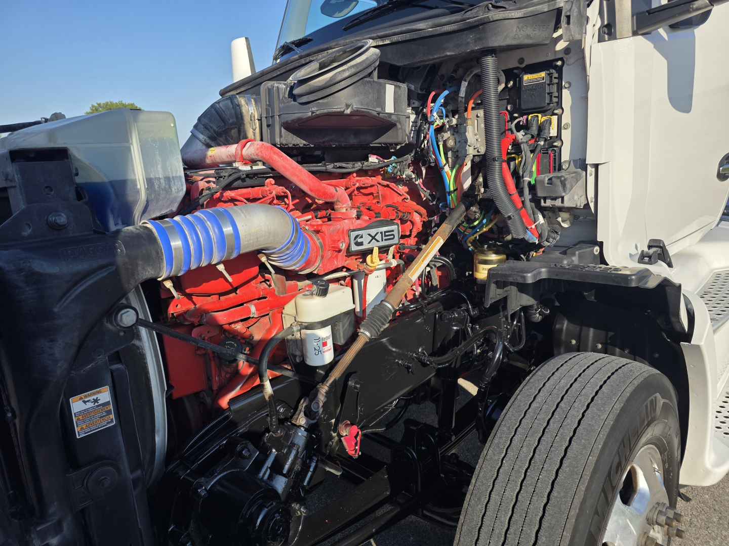 Kenworth T680 engine bay with red Cummins X15 engine visible, blue intake hose, wiring harnesses, and surrounding components.