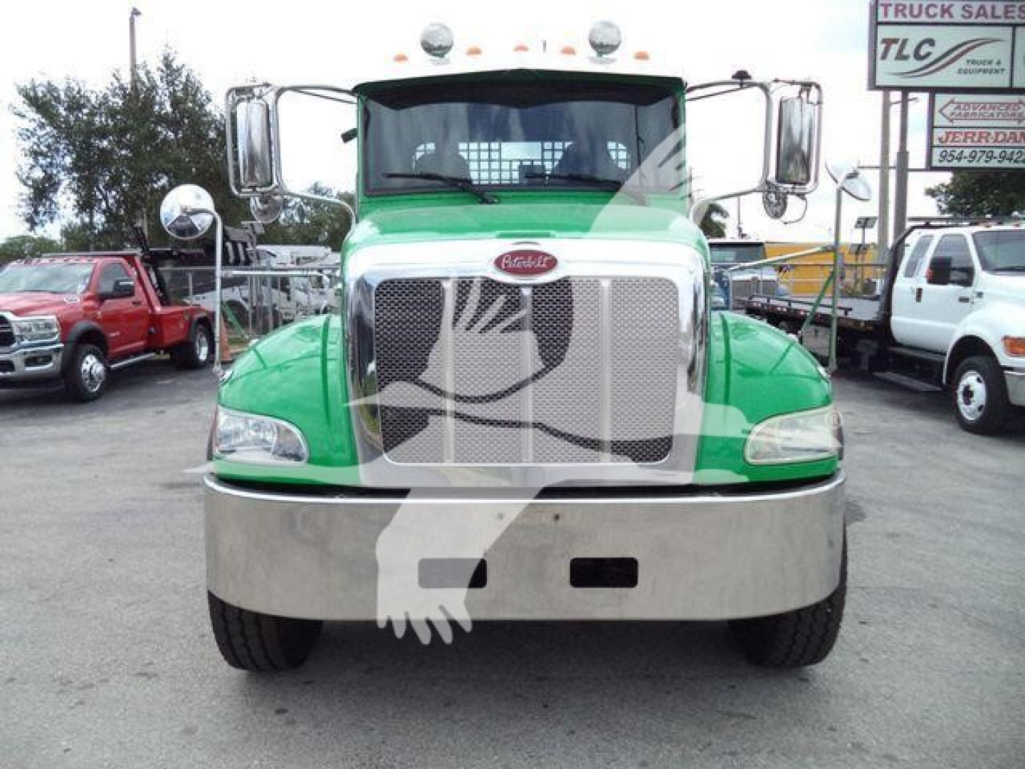 Front view of a green Peterbilt truck with a large chrome grille and bumper, used in a dealership lot. Visible text includes Peterbilt badge on grille, and a signboard in background reading 'TRUCK SALES' and contact numbers.