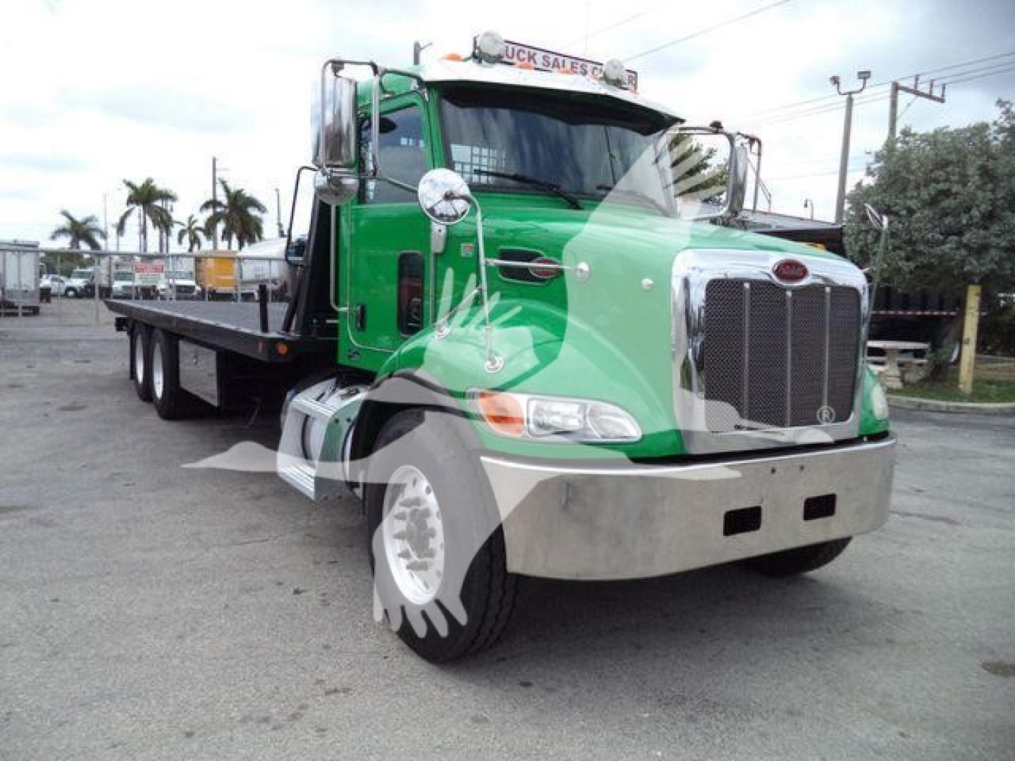 Peterbilt 348 rollback tow truck in green and chrome, with front grille, headlights, and long flatbed. Window reflection and dealership watermark overlaid.