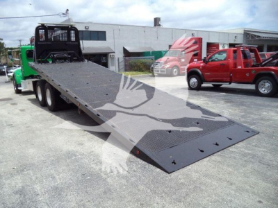 Peterbilt 348 rollback truck with a long loading ramp extended, dual rear axles, and a black mesh cab guard; office buildings and other trucks visible in background.